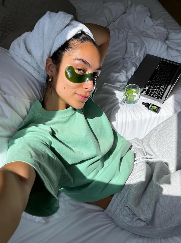 Person lying on a bed with green eye masks, wearing a towel, and using a laptop.